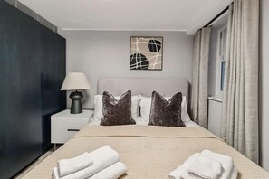 A hotel room with a bed, pillows, a nightstand, a lamp, a wall art piece, and curtains. - 7 Mins Walk to Selfridges Luxury Marble Arch Apt (London)