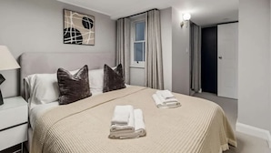 Habitación de hotel con cama, almohadas, mesita de noche, lámpara, ventana con cortinas, puerta y una obra de arte en la pared.