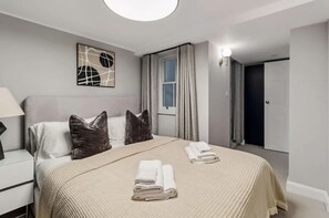 A hotel room with a bed, pillows, a nightstand, a lamp, a window with curtains, a door, and a wall art piece. - 7 Mins Walk to Selfridges Luxury Marble Arch Apt (London)