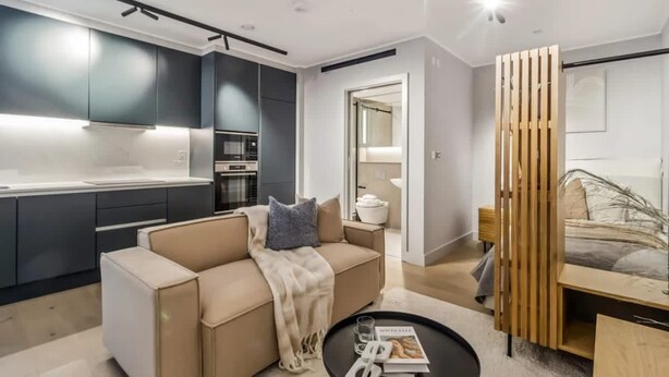 A modern living room with a sofa, a coffee table, and a kitchen area with cabinets and appliances. - 9 Mins Walk to Selfridges Marble Arch Studio (London)