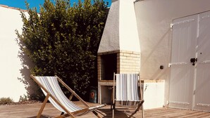 Una terraza de madera con dos sillones de jardín a rayas, una piscina pequeña y un edificio de ladrillo con chimenea.