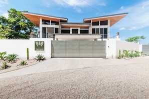 A modern two-story house with a flat roof, a large garage door, and a well-maintained garden with various plants. - Hilltop Villa: Infinity Pool & Ocean Horizon (Playa Grande)