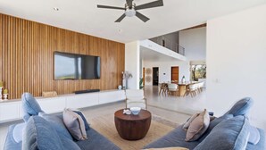 Uma sala de estar moderna com televisão de tela plana, painel de parede em madeira, ventilador de teto e uma área de refeições ao fundo.