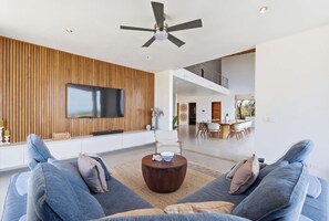 A modern living room with a flat-screen TV, wooden panel wall, ceiling fan, and a dining area in the background. - Hilltop Villa: Infinity Pool & Ocean Horizon (Playa Grande)