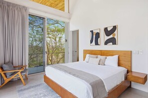A modern bedroom with a large bed, a wooden headboard, a nightstand, and a view through a sliding glass door to an outdoor balcony. - Hilltop Villa: Infinity Pool & Ocean Horizon (Playa Grande)