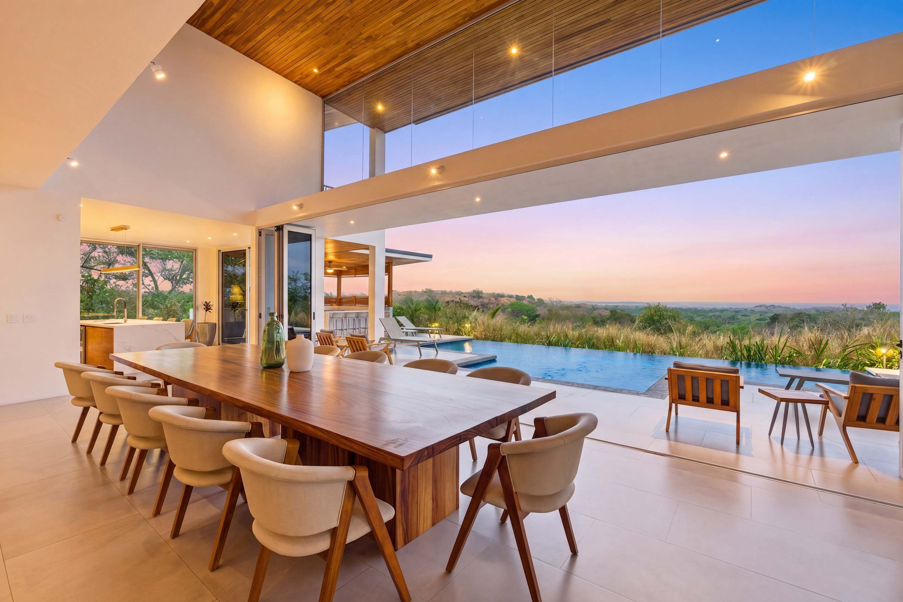 A modern dining area with a large wooden table and chairs, a pool, and a view of the landscape.