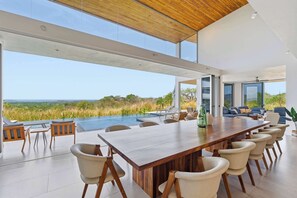 A modern dining area with a long wooden table, comfortable chairs, and a view of the landscape. - Hilltop Villa: Infinity Pool & Ocean Horizon (Playa Grande)