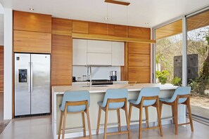 A modern kitchen with wooden cabinets, a stainless steel refrigerator, and a bar area with stools. - Hilltop Villa: Infinity Pool & Ocean Horizon (Playa Grande)