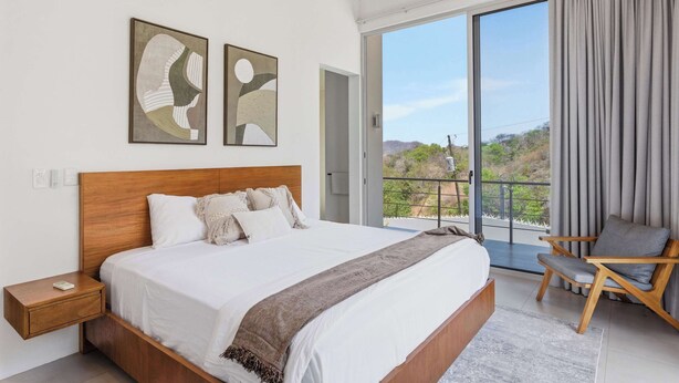 Quarto com uma cama grande, cabeceira de madeira, mesa de cabeceira, uma cadeira e varanda com vista.