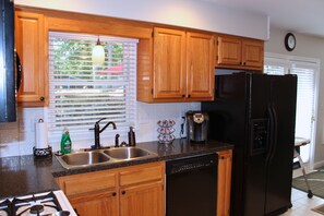 A kitchen with wooden cabinets, a black refrigerator, a coffee maker, and a window with blinds. - 30+ Day Stay  Fully Furnished 4BR Home Near Sentara RMH & JMU (Harrisonburg)