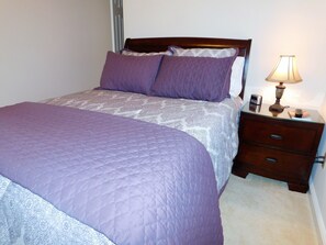Ein Bett mit einer gesteppten, violetten Decke, einem gemusterten Überwurf und zwei Kissen. Ein Nachttisch mit Lampe, einer Uhr und einem Buch sind ebenfalls vorhanden.