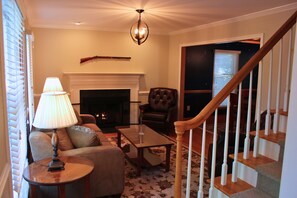 A living room with a fireplace, a sofa, a coffee table, a lamp, and a staircase. - 30+ Day Stay  Fully Furnished 4BR Home Near Sentara RMH & JMU (Harrisonburg)