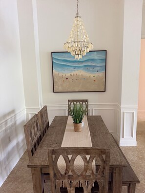 A dining area with a wooden table, chairs, a hanging chandelier, and a beach-themed painting on the wall. - Resort Style Living. Beautiful Coastal Inspired Coach Home in Fiddler’s Creek. (Naples)