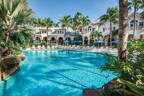 Ein Pool mit Liegestühlen und Sonnenschirmen, umgeben von Palmen und einem Gebäude mit Bögen.