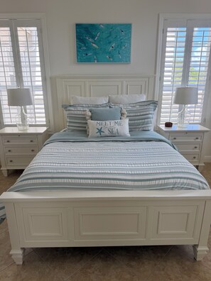 A bed with a striped bedspread, two bedside tables with lamps, a wall-mounted painting of swimmers, and a pillow with text. - Resort Style Living. Beautiful Coastal Inspired Coach Home in Fiddler’s Creek. (Naples)