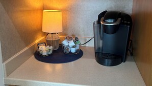 Ett litet kök med träskåp, en kaffebryggare, en lampa och en korg med kaffekapslar.
