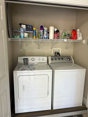 A laundry room with two white washing machines, a shelf above containing various cleaning products, and a power outlet on the wall. - Lakefront Paradise on Lake Hickory’s Main Channel – Handicap Accessible (Taylorsville)
