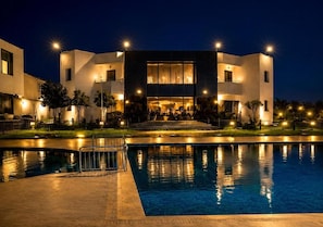 A modern hotel with a swimming pool, a covered outdoor area, and a well-lit entrance.