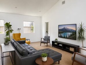 Ein modernes Wohnzimmer mit einem Ecksofa, einem gelben Sessel, einem grauen Sessel, einem Couchtisch und einem großen Fernseher auf einem schwarzen Lowboard.