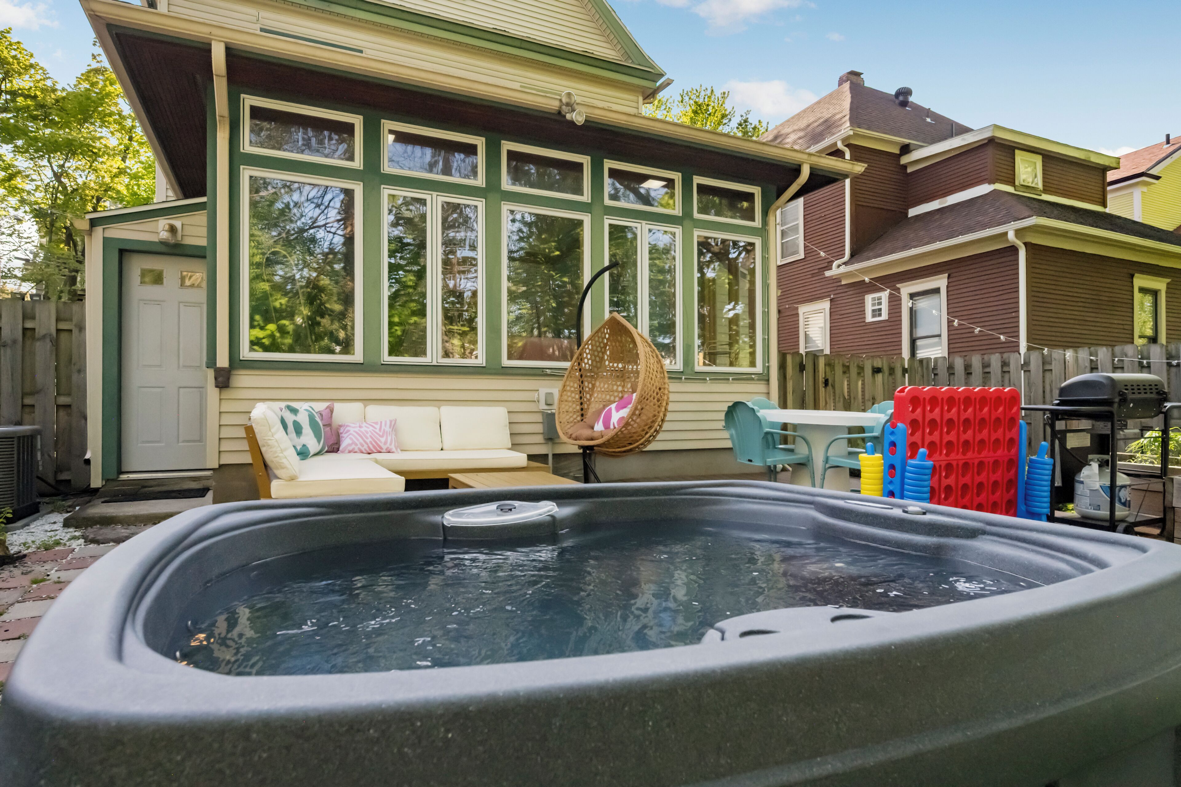 A hot tub in a backyard with a wooden fence, a white door, and a beige house with green trim.