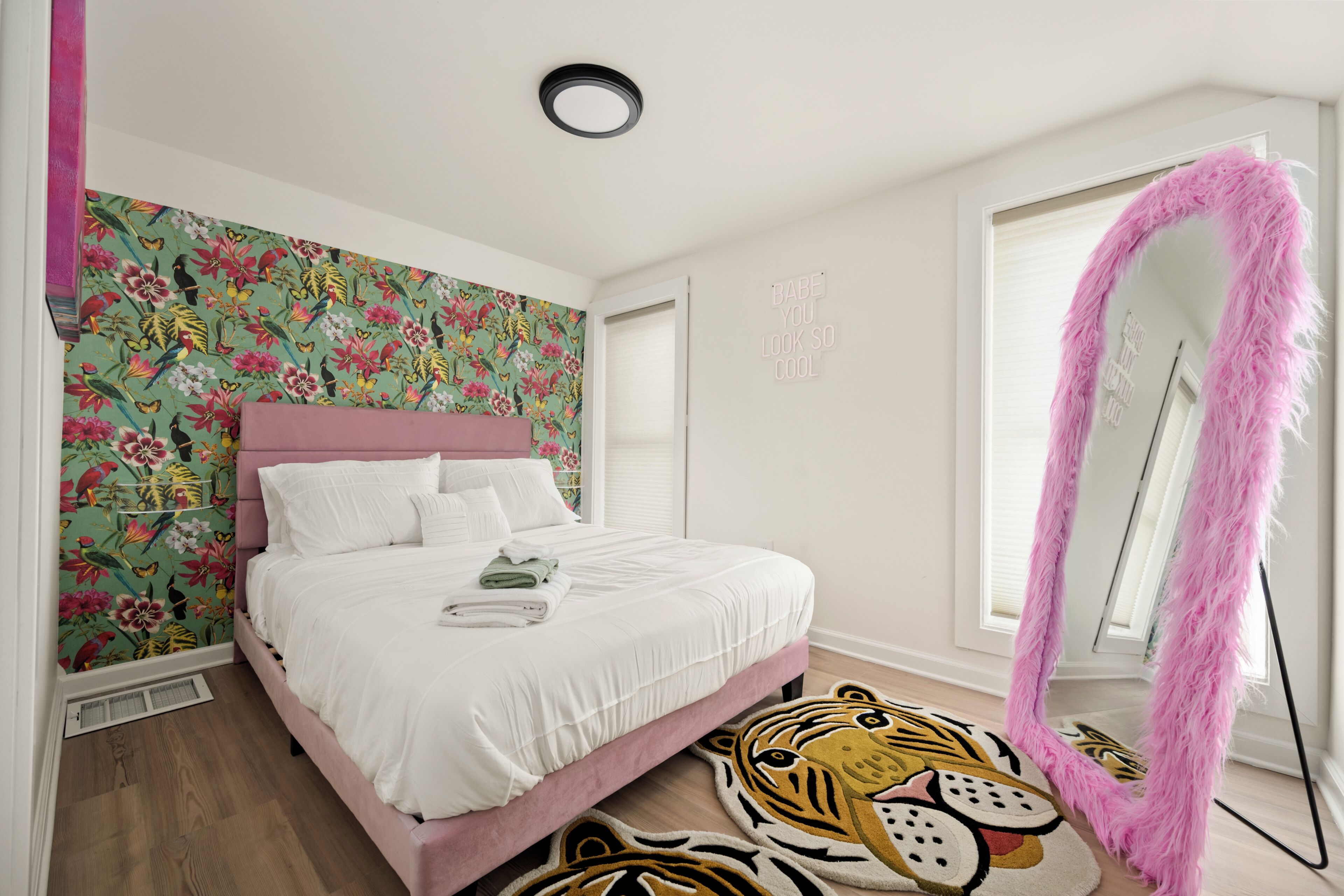 A bedroom with a floral wallpaper, a pink bed, a tiger rug, and a mirror with a pink frame.