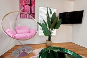 Een kamer met een hangstoel, een kamerplant, een televisie en een raam met jaloezieën.