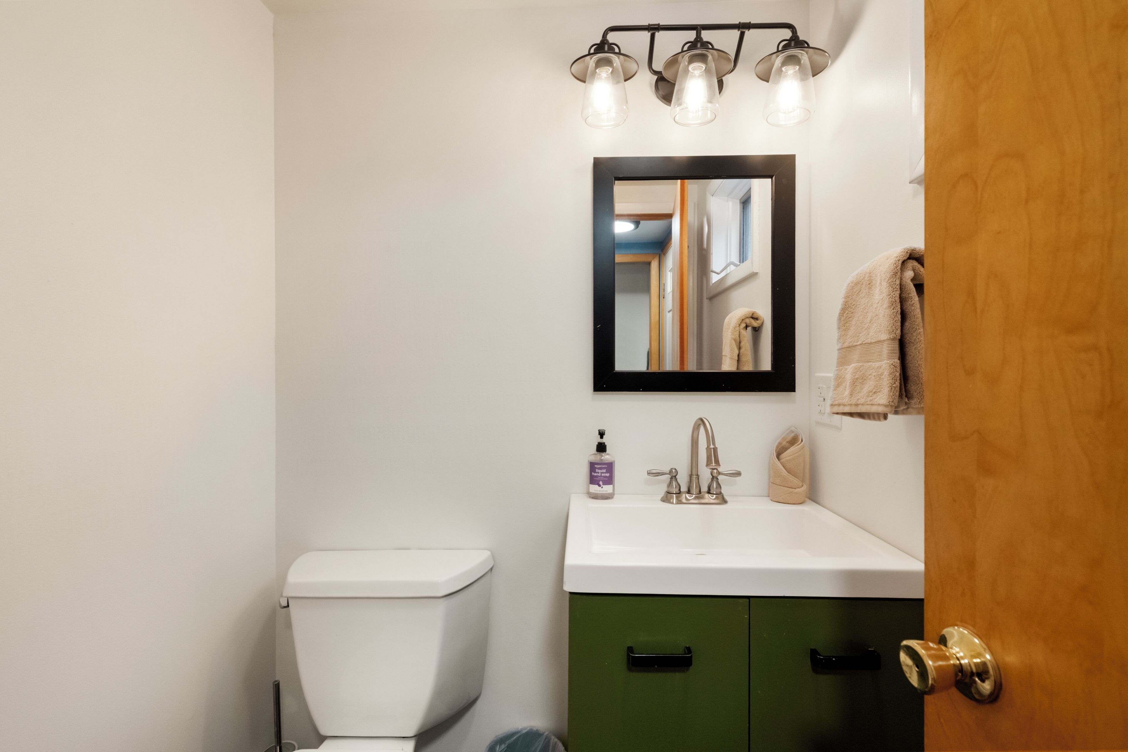 Une salle de bain comprenant des toilettes, un lavabo, un meuble vert et un miroir avec des lumières au-dessus.