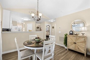 A dining area with a round table, white chairs, a chandelier, and a wooden cabinet with a mirror. - Coastal Chic Golf Retreat Fairways 18F (Myrtle Beach)