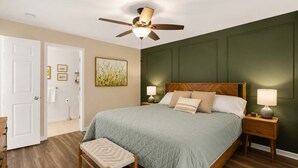 Ein Schlafzimmer mit einem Bett, einem Nachttisch, einer Nachttischlampe, einem Deckenventilator, einer Tür und einem Wandbild.