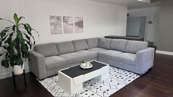 Una sala moderna con un sofá seccional gris, una mesa de centro blanca, una planta grande en maceta y una alfombra estampada.