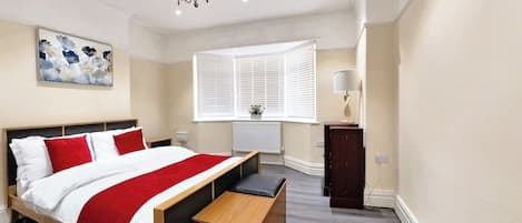 A bedroom with a bed, bedside table, bench, window with blinds, radiator, and a chandelier.