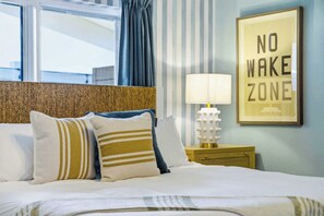 Ein Hotelzimmer mit Bett, Kissen, einem Nachttisch mit Lampe, einem gerahmten Schild und einem Fenster mit Vorhängen.