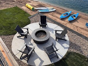 A circular outdoor seating area with a fire pit, surrounded by chairs, near a beach with kayaks. - Sugar Sand Sunset | Beachfront | Sleeps 20 (New Lisbon)