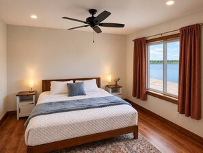 Una camera da letto con un letto, una ventola a soffitto, lampade da comodino, una finestra con tende e vista su un corpo d'acqua.
