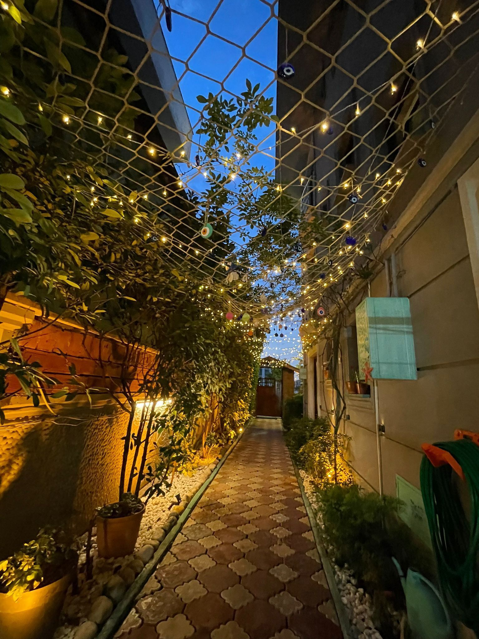 A narrow pathway with a tiled floor, surrounded by greenery and decorative lights.