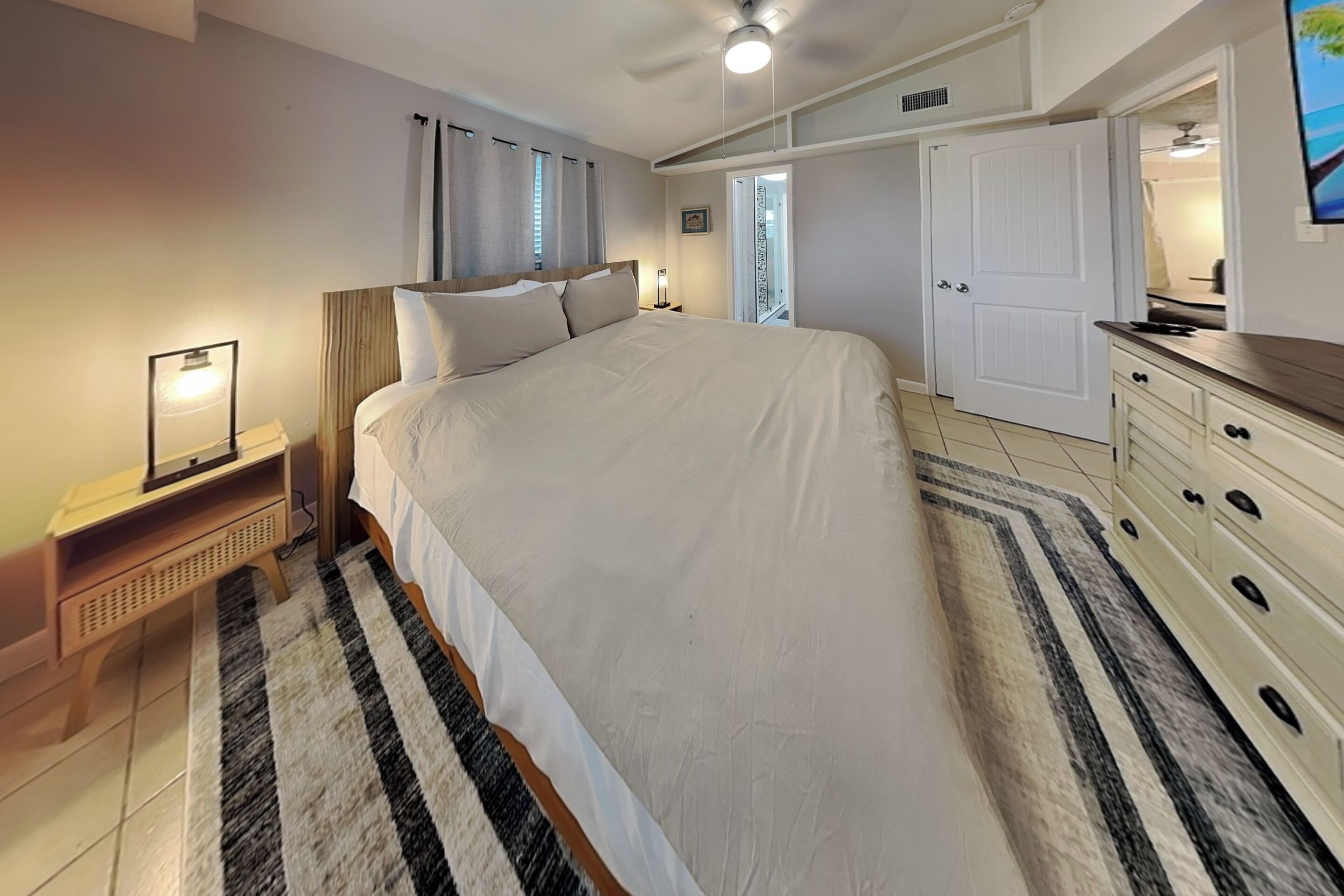 A bedroom with a large bed, a bedside table with a lamp, a dresser, and a door leading to a bathroom.