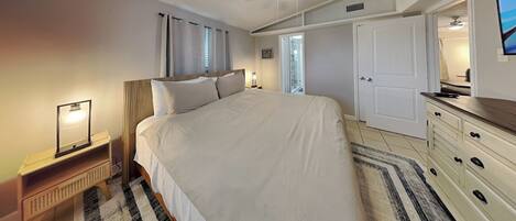 A bedroom with a large bed, a bedside table with a lamp, a dresser, and a door leading to a bathroom.