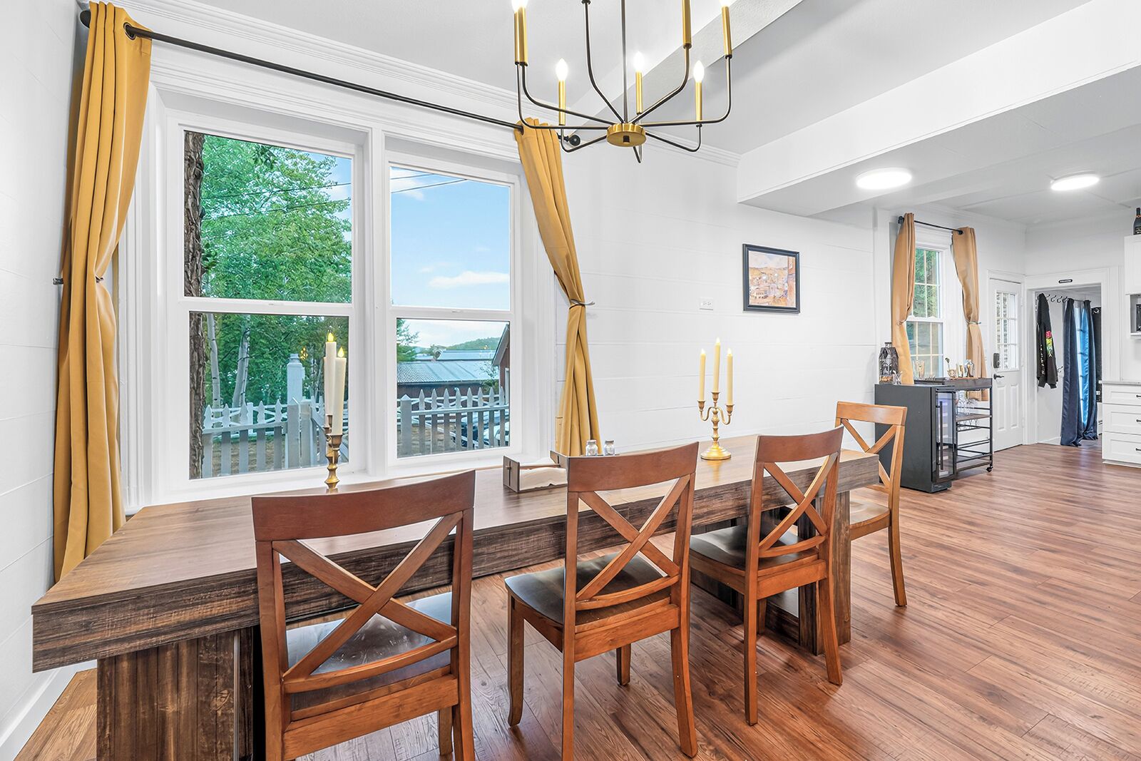 A dining area with a wooden table and chairs, large windows with curtains, and a chandelier.