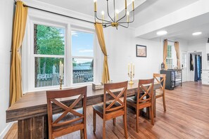 A dining area with a wooden table and chairs, large windows with curtains, and a chandelier.