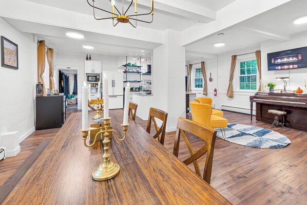 A modern kitchen with white cabinets, a dining area with a wooden table and chairs, and a living room with a piano and a television.