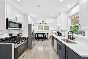 A modern kitchen with white cabinetry, stainless steel appliances, a central island with bar stools, and a window providing a view of greenery. - Creek Retreat on 1 acre / Game Room / Fire Pit (Matteson)