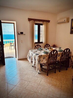 A dining area with a view of the sea, a window with curtains, and a tiled floor. - Villa Mousouna - Panoramic Sea View of Lefkos (Kato Lefkos)