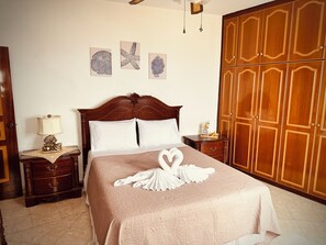 A neatly made bed with a heart-shaped towel arrangement, a wooden nightstand with a lamp, and a large wardrobe. - Villa Mousouna - Panoramic Sea View of Lefkos (Kato Lefkos)