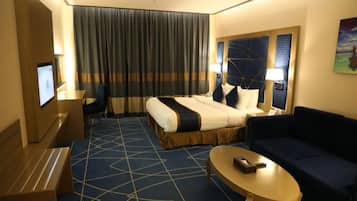 Habitación de hotel con cama, sofá, televisión, escritorio y un ventanal con cortinas.
