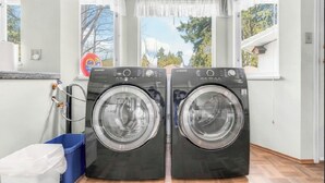 A laundry room with two front-loading washing machines, a blue laundry basket, and a white trash bin.