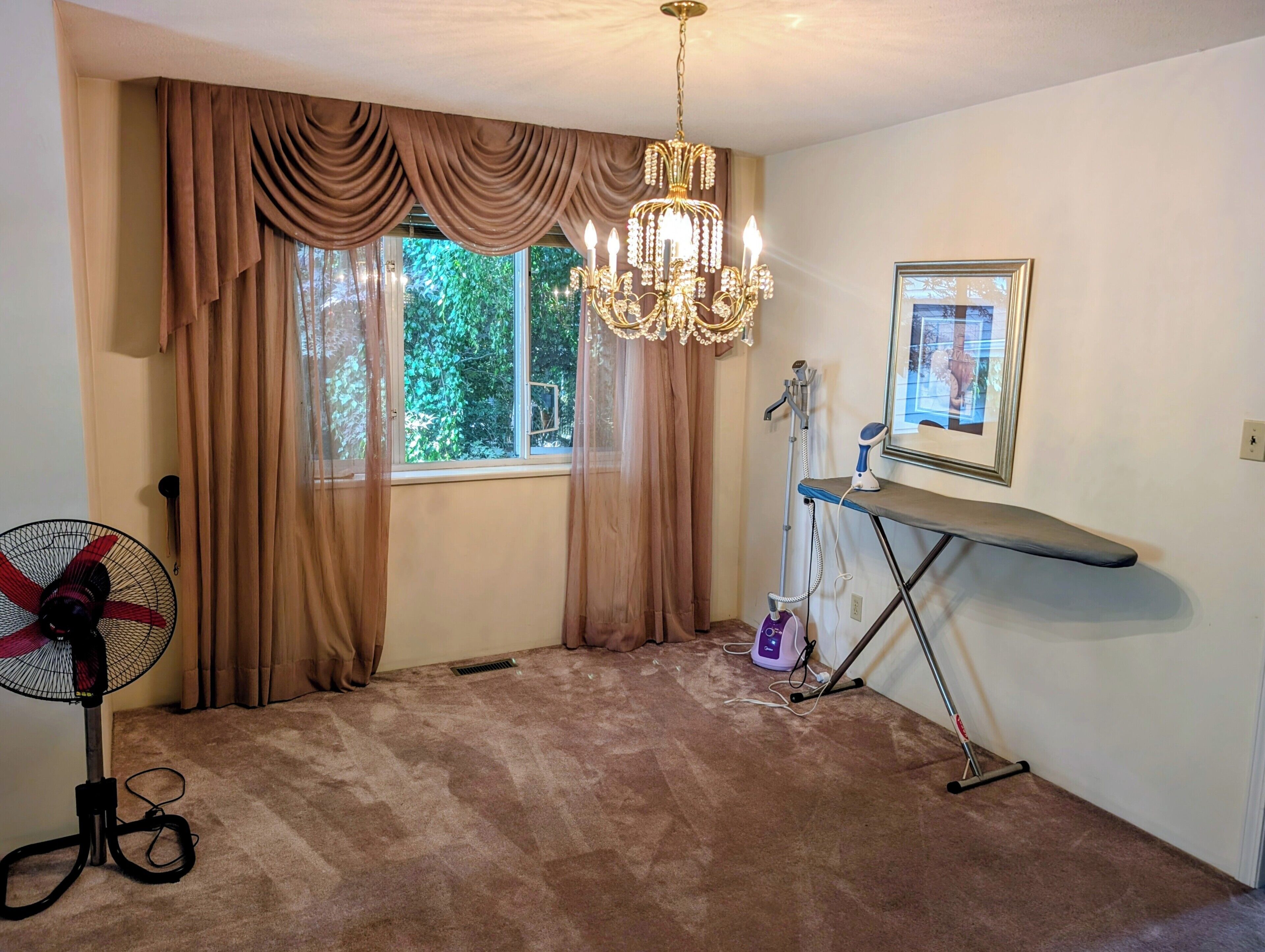 A room with a large window, a standing fan, a vacuum cleaner, and a framed picture on the wall.