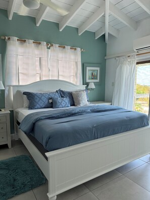 A bedroom with a white bed, blue bedding, a teal rug, and a window with curtains.