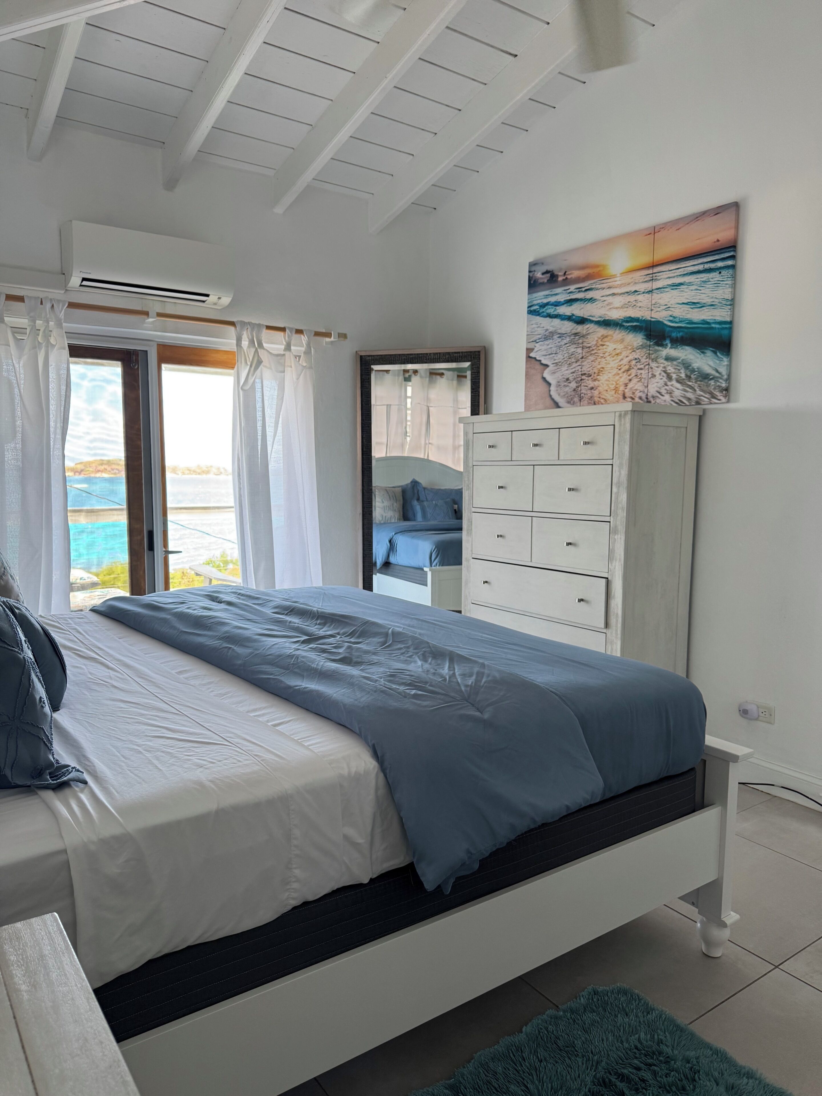 A bedroom with a bed, a dresser, a painting, and a view of the sea through a window.