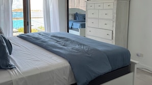 Een slaapkamer met een bed, een kaptafel, een schilderij en uitzicht op zee door een raam.