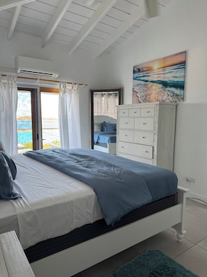 Ein Schlafzimmer mit einem Bett, einer Kommode, einem Bild und Meerblick durch ein Fenster.